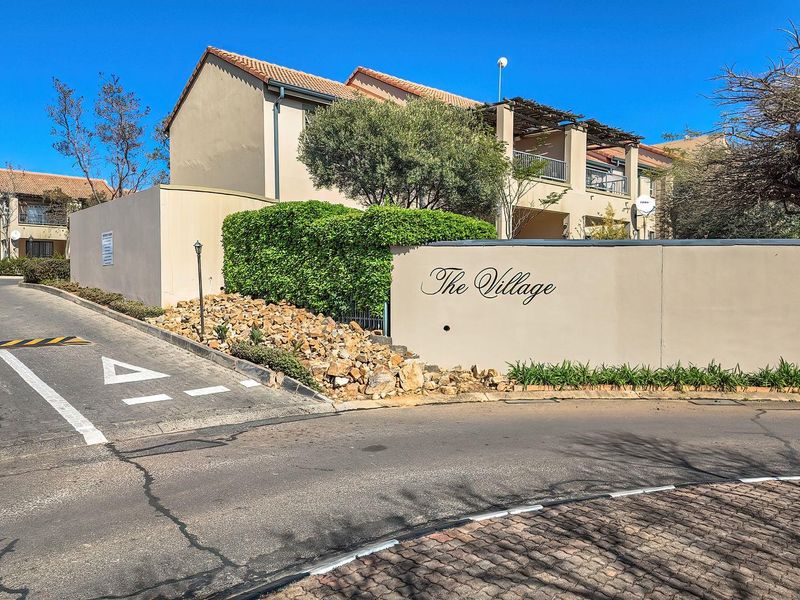 Street view of the entrance to The Village apartment complex, featuring a paved driveway, boundary wall, and multi-level residential units with balconies.