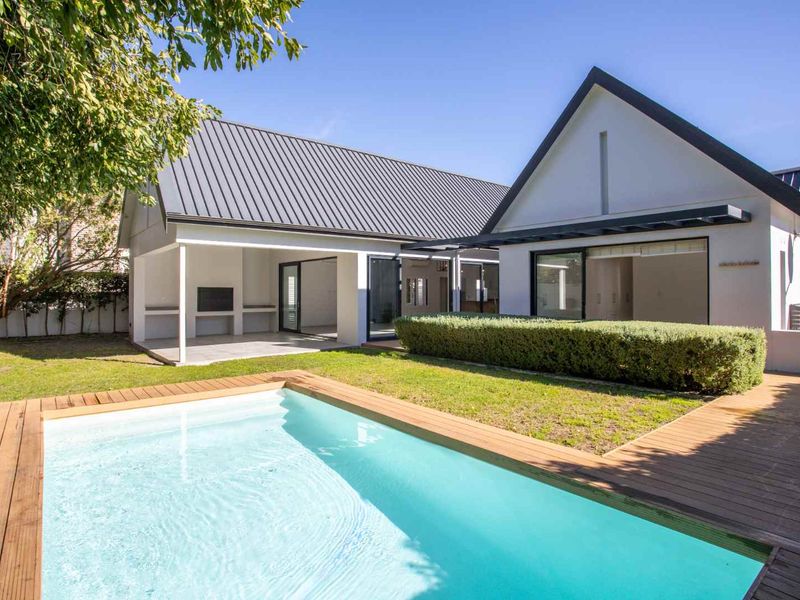 Rear view of the property featuring a modern swimming pool, timber decking, a covered patio with a built-in braai, and well-maintained garden space.