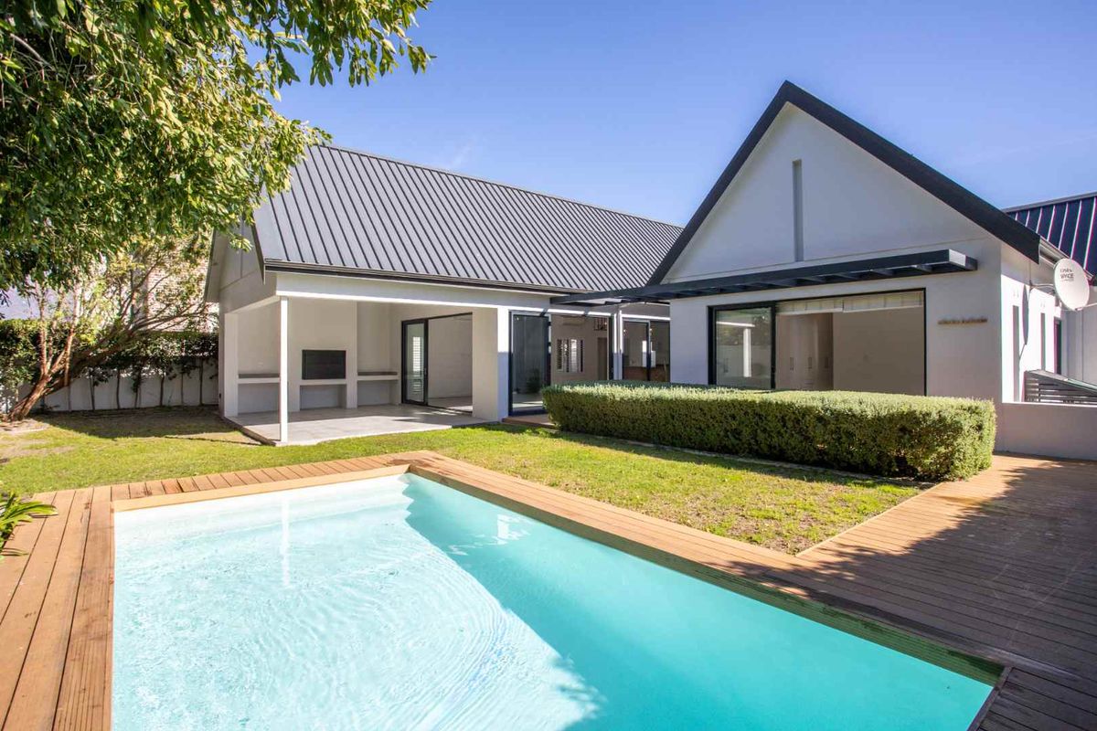 Rear view of the property featuring a modern swimming pool, timber decking, a covered patio with a built-in braai, and well-maintained garden space.