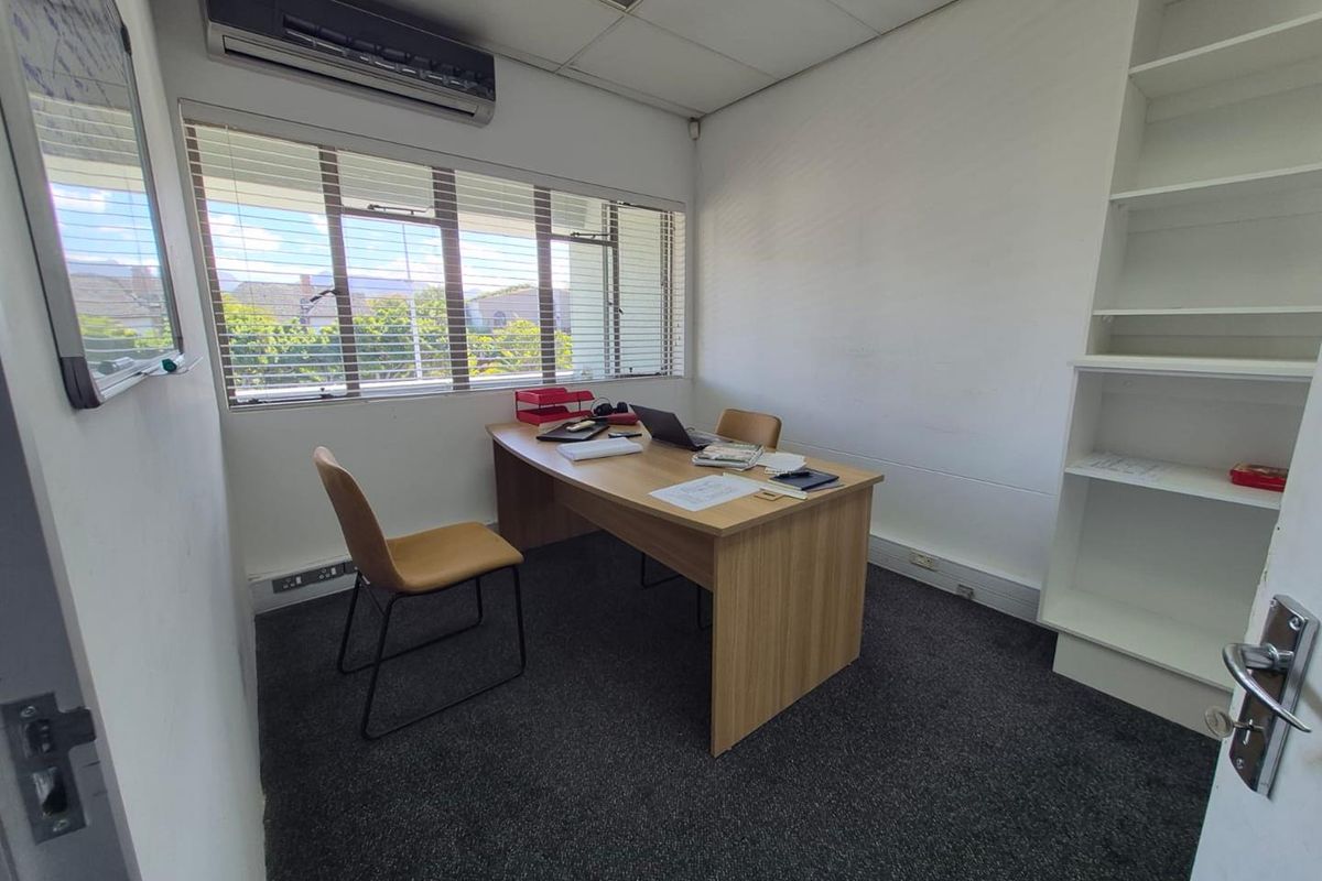 A functional office space featuring carpeted flooring, built-in shelving, an air conditioning unit, and large windows with venetian blinds.