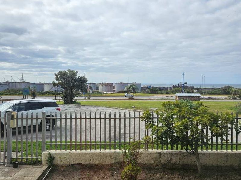 An exterior view showing a metal perimeter fence, a retaining wall, and a paved walkway overlooking an industrial area and distant coastline.