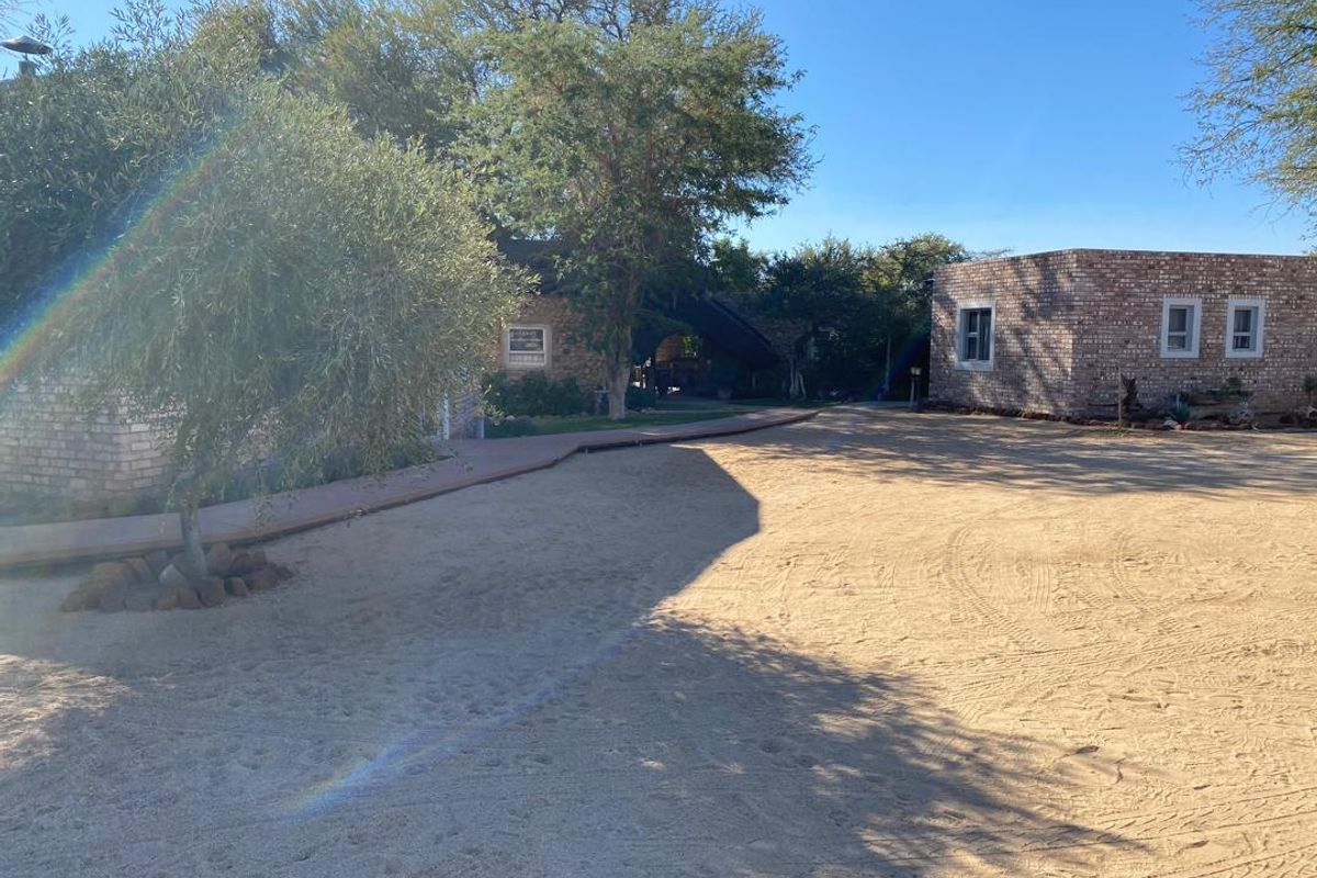 A view of the rear exterior area featuring exposed brick buildings, a wooden walkway, and mature trees set within a sandy landscape.
