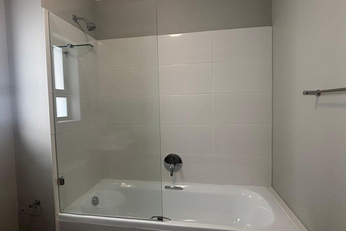 A clean, modern bathroom featuring a built-in bathtub with a glass shower screen, chrome fixtures, and white wall tiling.