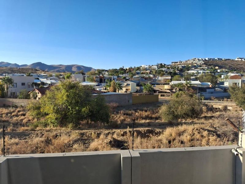 An elevated view overlooking a residential neighborhood and surrounding hills, featuring a concrete perimeter wall with electric fencing in the foreground.