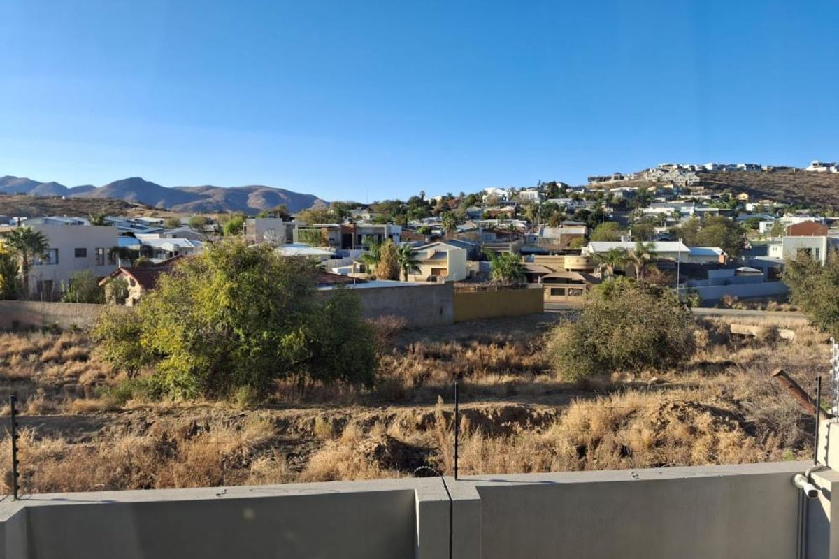 An elevated view overlooking a residential neighborhood and surrounding hills, featuring a concrete perimeter wall with electric fencing in the foreground.