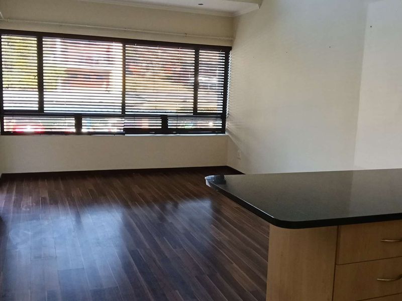 An open-plan living area featuring dark laminate flooring, a kitchen island with built-in cabinetry, and large windows fitted with venetian blinds.