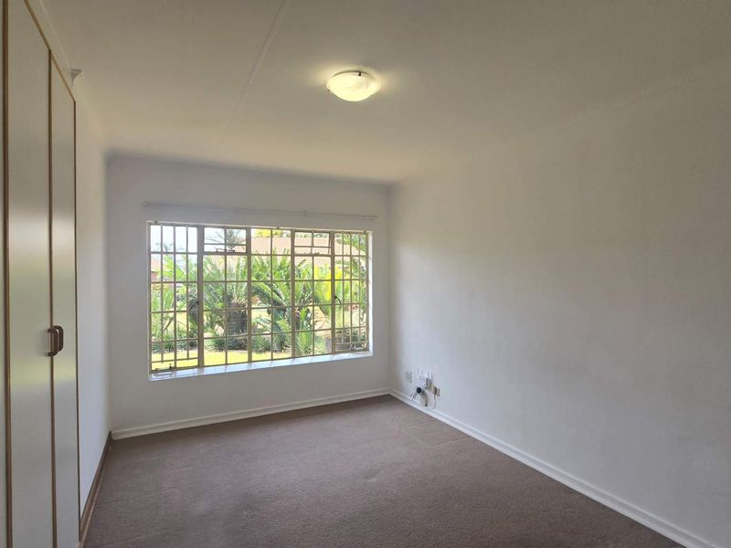 A bright, empty bedroom featuring built-in cupboards, carpeted flooring, and a large window with security bars overlooking the garden.