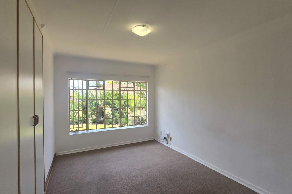 A bright, empty bedroom featuring built-in cupboards, carpeted flooring, and a large window with security bars overlooking the garden.