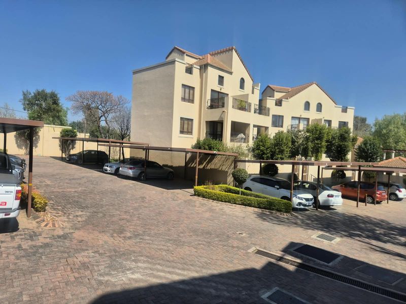 Exterior view of an apartment complex featuring a paved parking area with covered carports and landscaped garden beds.