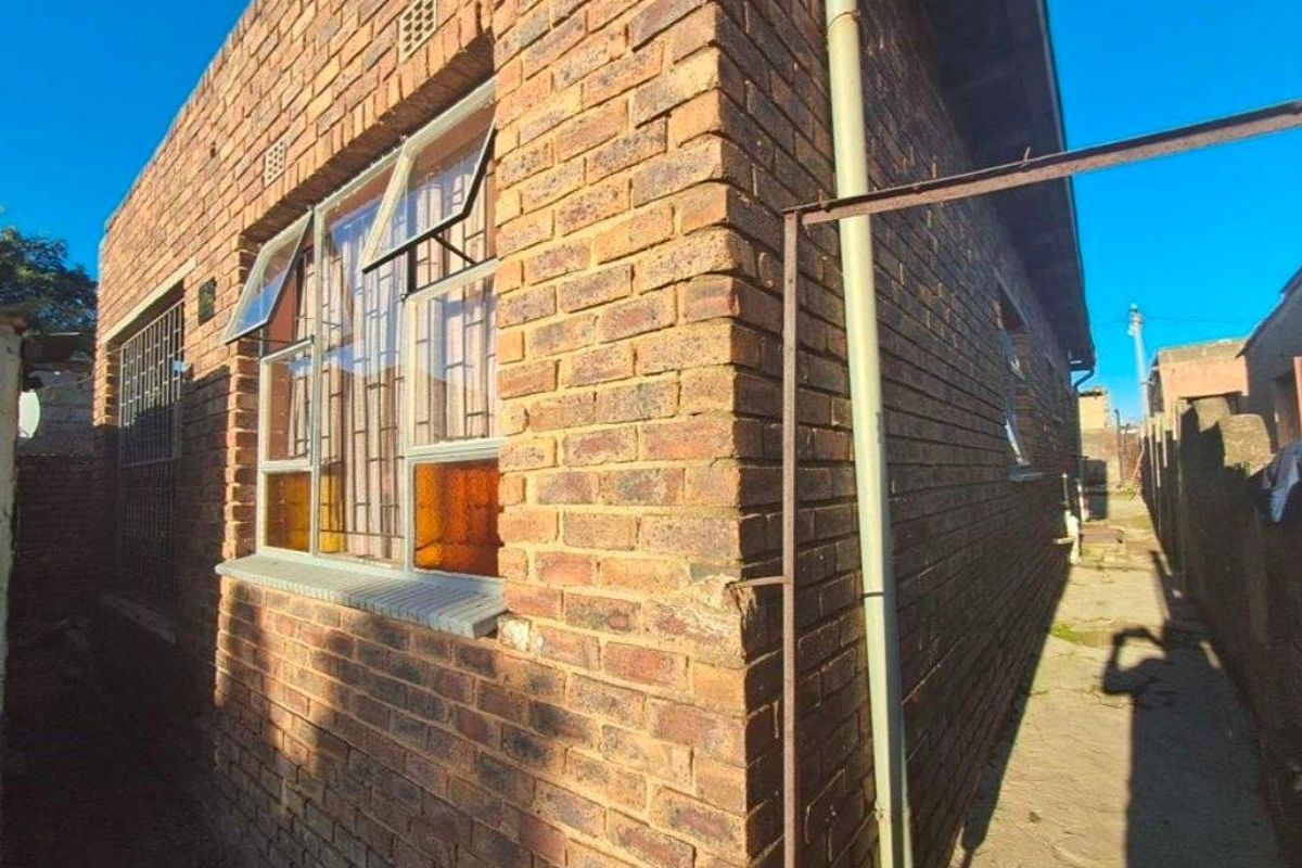 Side view of the house featuring exposed face-brick exterior walls, metal-framed windows with security bars, and a narrow concrete pathway.