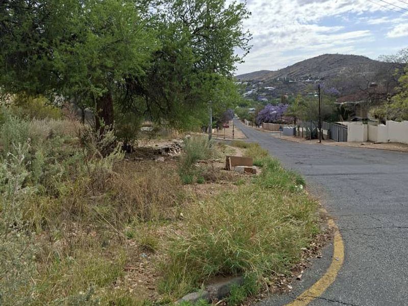 A street view showing the asphalt road, concrete curbing, and surrounding landscape with overhead power lines and street lighting in a residential area.