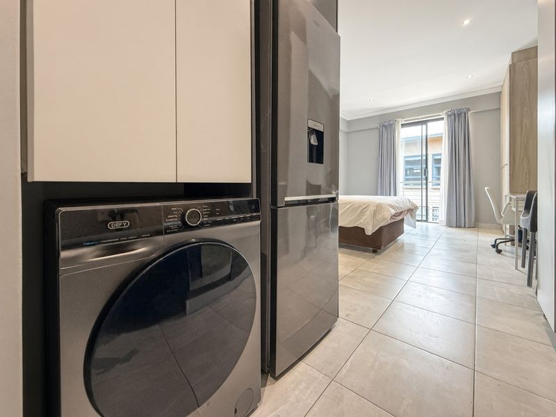 An open-plan living space featuring tiled flooring, recessed lighting, and integrated cabinetry with a laundry area, leading to a balcony through sliding glass doors.