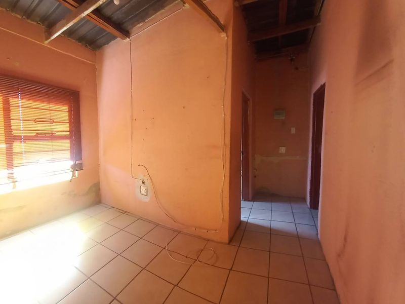 An empty living area featuring tiled floors, exposed timber ceiling beams, and venetian blinds, requiring cosmetic renovation and wall repairs.