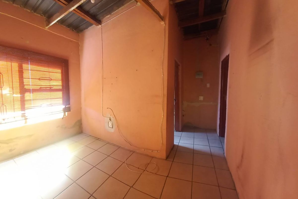 An empty living area featuring tiled floors, exposed timber ceiling beams, and venetian blinds, requiring cosmetic renovation and wall repairs.