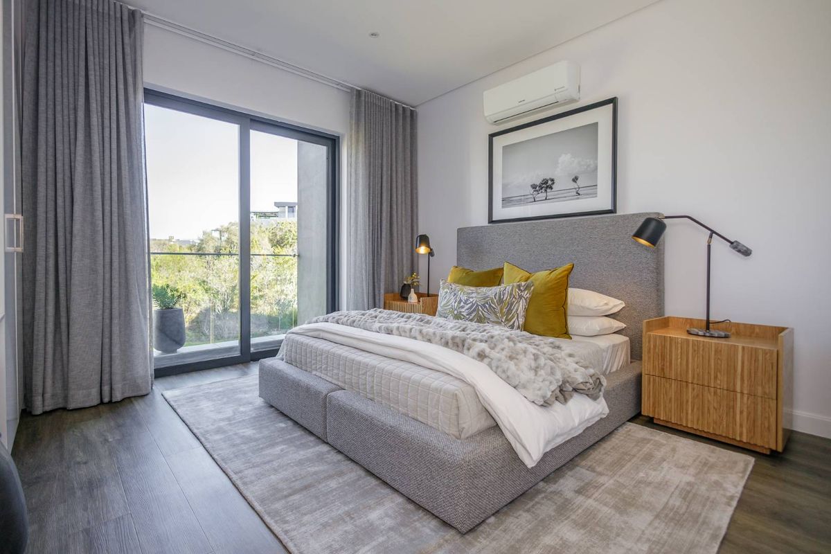 A modern, well-lit bedroom featuring wood-look flooring, a wall-mounted air conditioning unit, and large sliding glass doors leading to a private balcony.