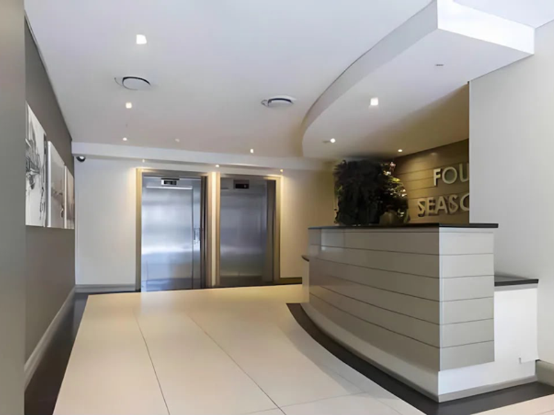 Modern apartment building lobby featuring a sleek reception desk, stainless steel elevators, and contemporary recessed lighting with polished tiled flooring.