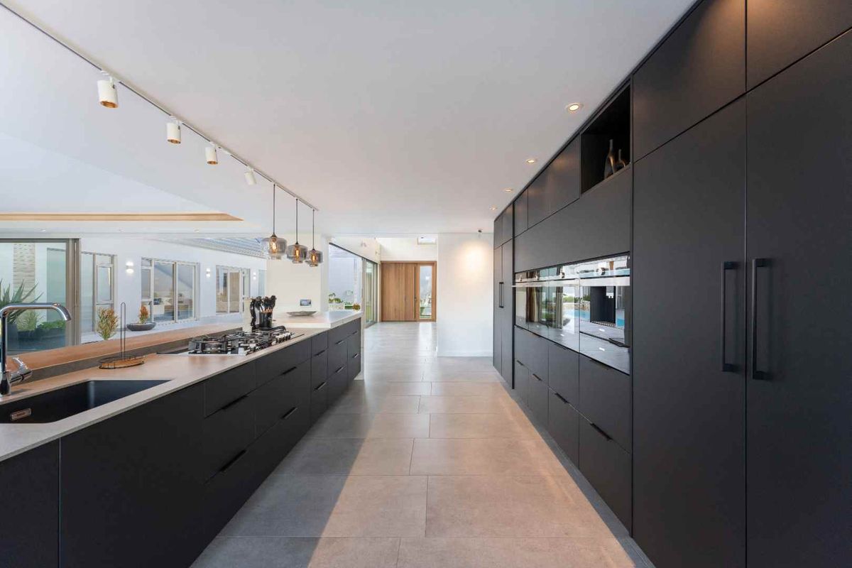 Modern, high-end kitchen featuring sleek matte black cabinetry, integrated appliances, stone countertops, and professional track lighting in an open-plan layout.