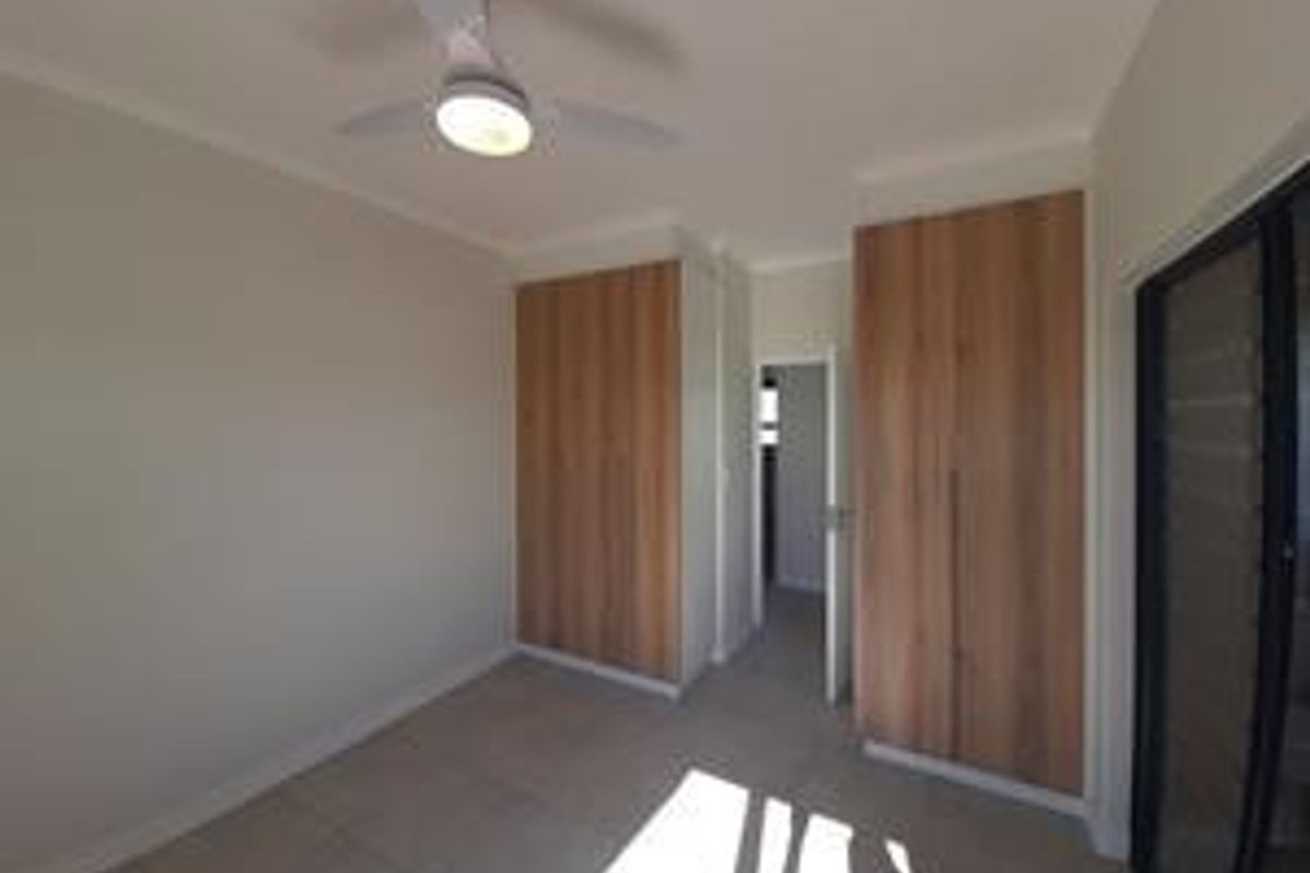 A bright, empty bedroom featuring modern wood-finish built-in cupboards, a ceiling fan, and neutral tiled flooring.