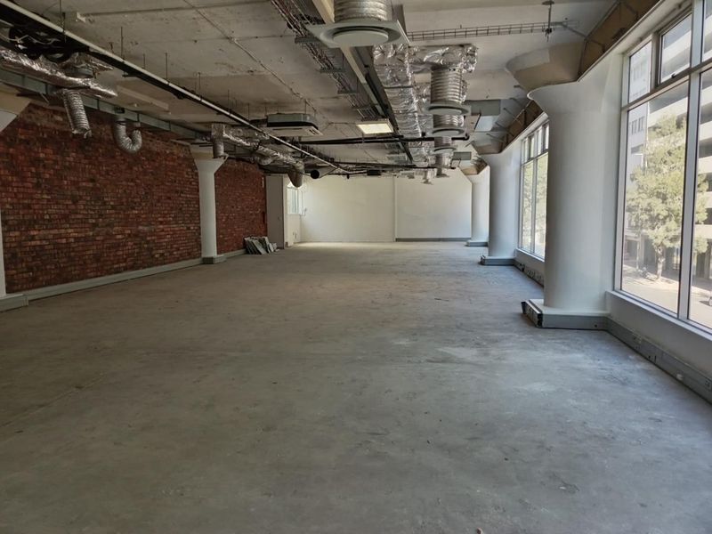 An open-plan office space featuring exposed brick walls, industrial-style exposed ceiling ducting, and large windows providing ample natural light.