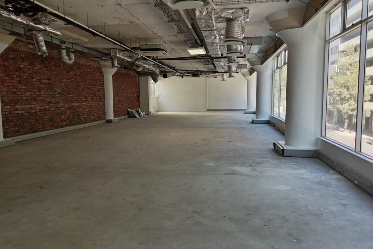 An open-plan office space featuring exposed brick walls, industrial-style exposed ceiling ducting, and large windows providing ample natural light.