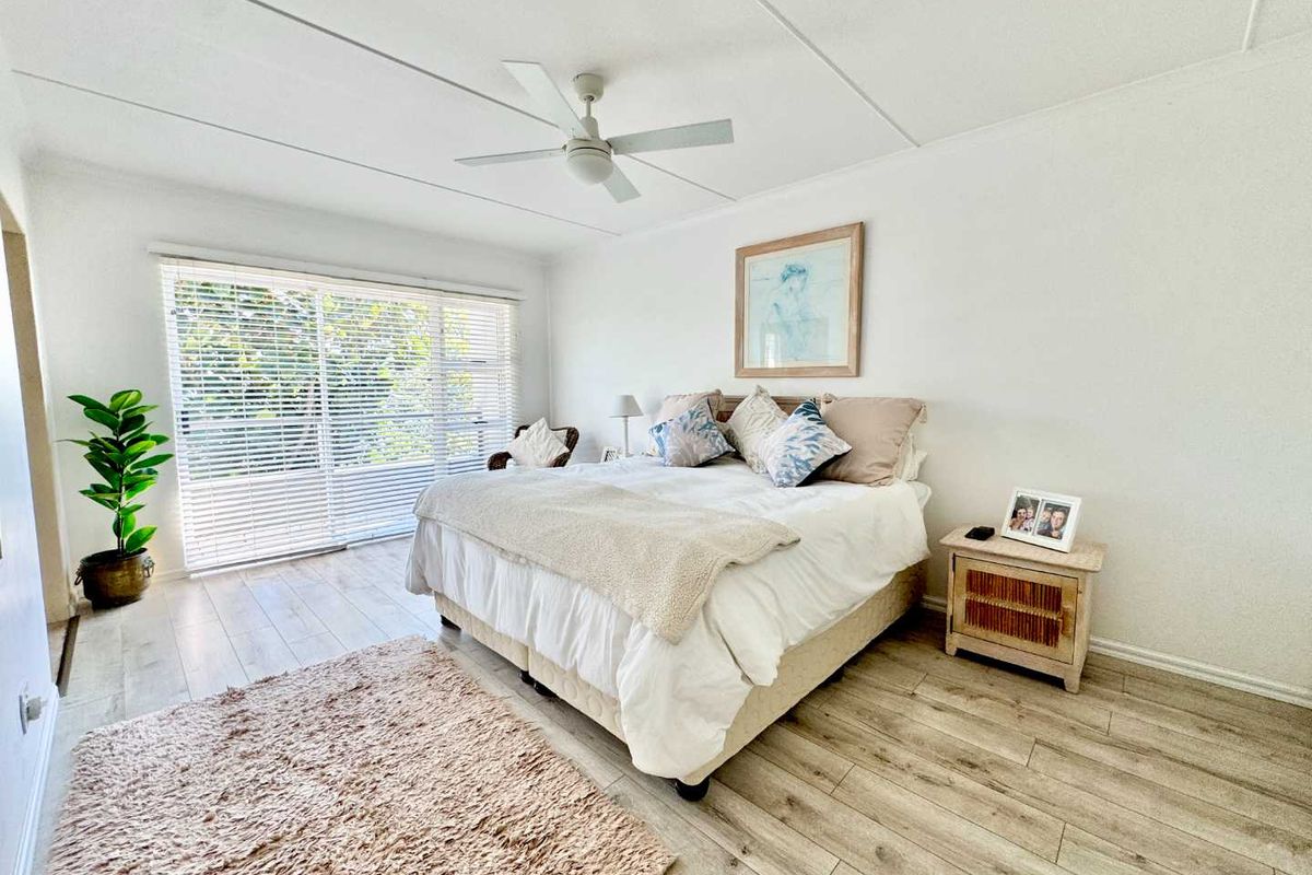 A bright and airy bedroom featuring light-toned laminate flooring, a ceiling fan, and large sliding glass doors with venetian blinds for natural light.