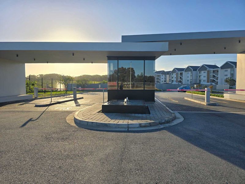 Modern security gatehouse entrance to the apartment complex featuring a central water feature, boom gates, and paved driveway access.