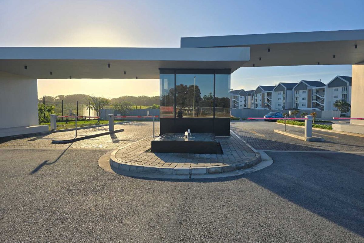 Modern security gatehouse entrance to the apartment complex featuring a central water feature, boom gates, and paved driveway access.