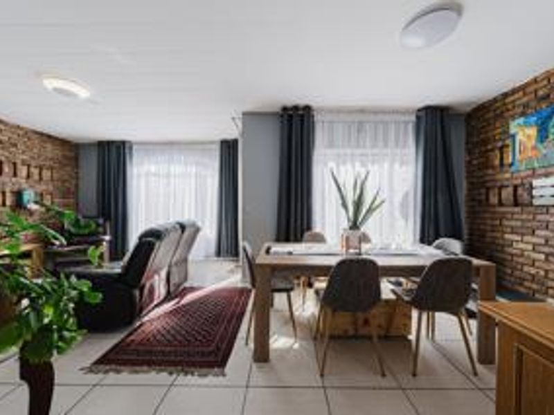 Spacious open-plan living and dining area featuring distinctive exposed brick walls, neutral tiled flooring, and ample natural light through large windows.
