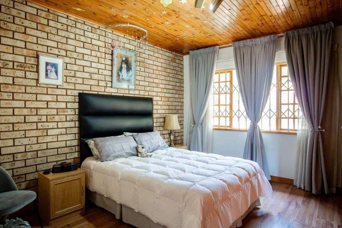 A bedroom featuring an exposed brick feature wall, timber ceiling, laminate flooring, and built-in cupboards with security bars on the windows.