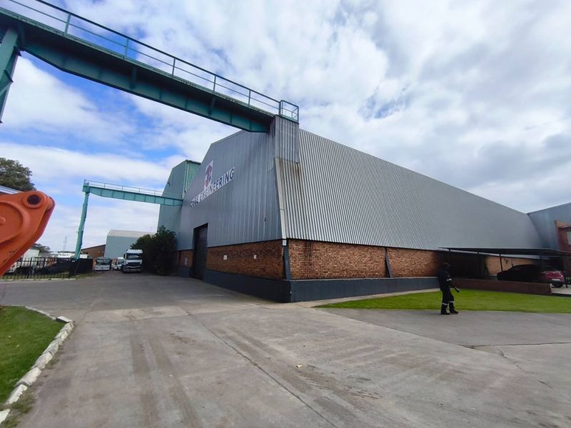 Exterior view of an industrial warehouse facility featuring corrugated metal cladding, a brick base, and an elevated steel walkway connecting structures.