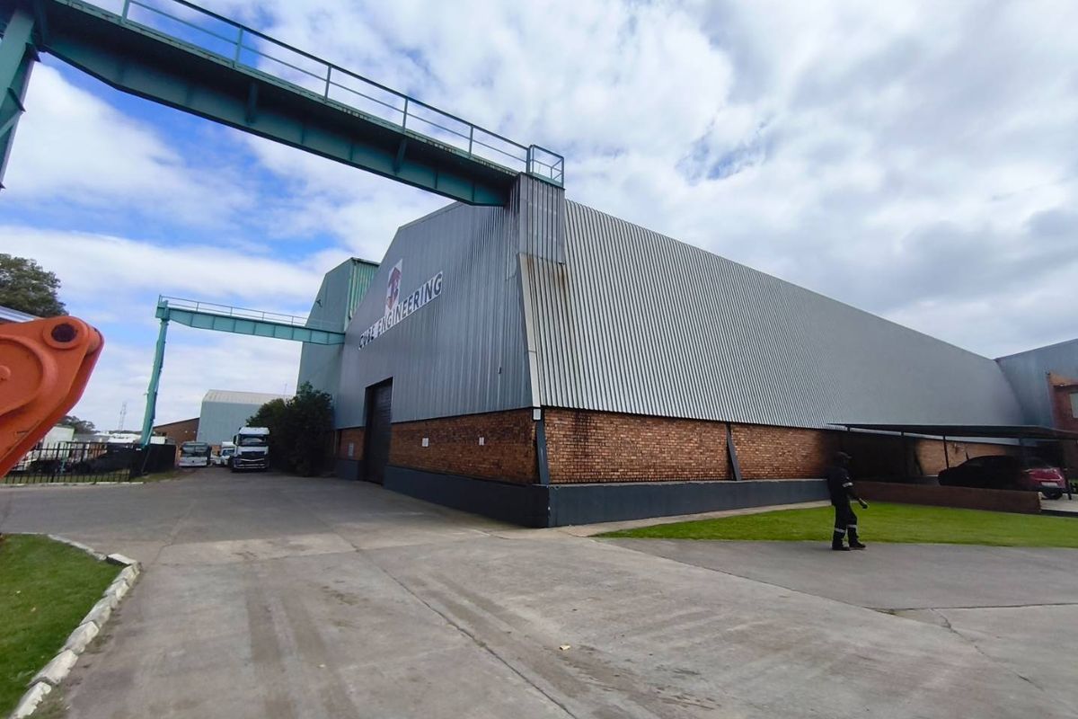 Exterior view of an industrial warehouse facility featuring corrugated metal cladding, a brick base, and an elevated steel walkway connecting structures.