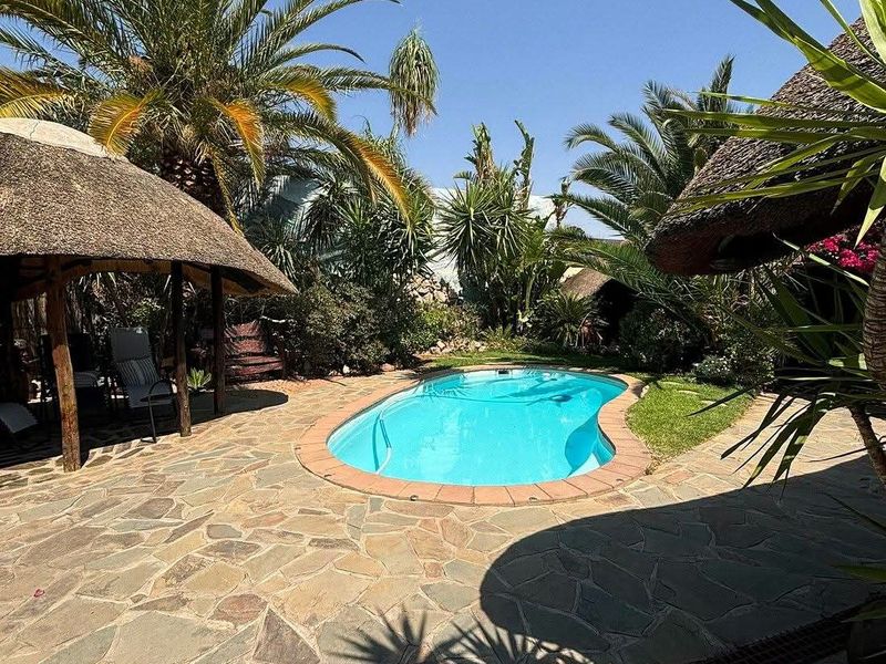A well-maintained outdoor pool area featuring a swimming pool, stone paving, and a traditional thatched lapa, surrounded by lush tropical landscaping.