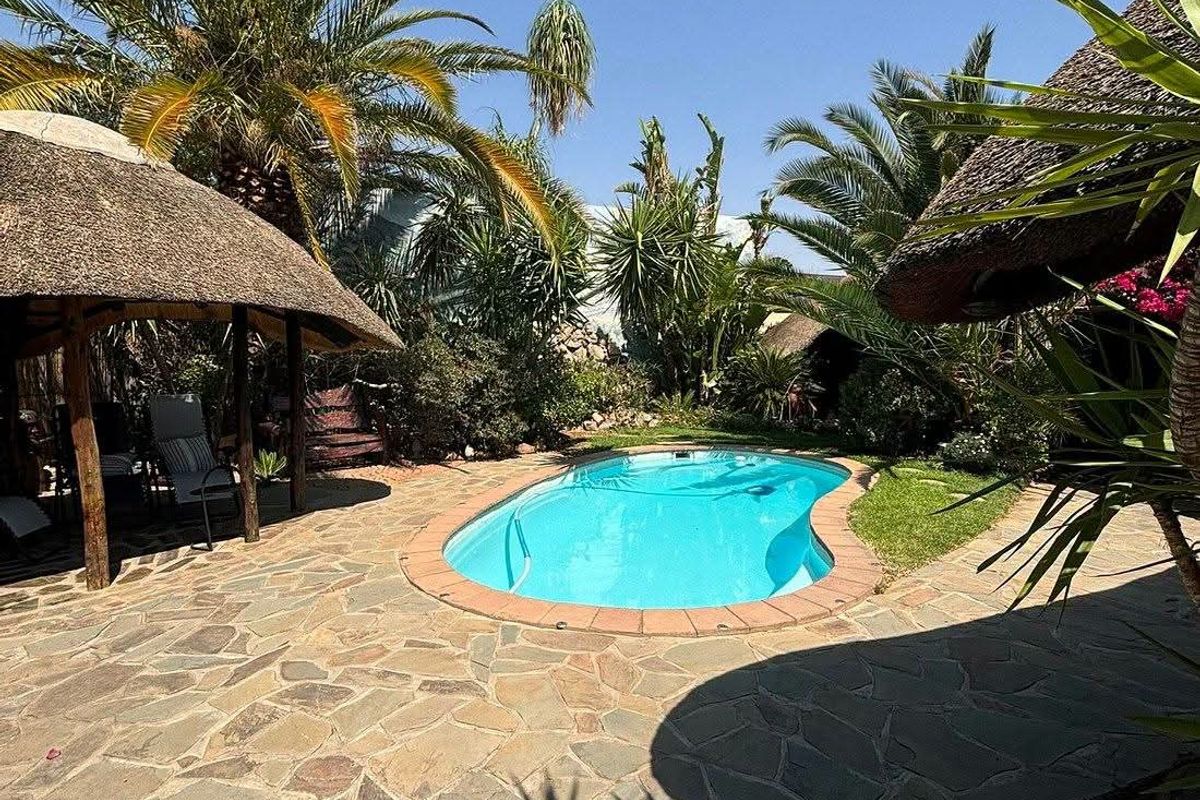 A well-maintained outdoor pool area featuring a swimming pool, stone paving, and a traditional thatched lapa, surrounded by lush tropical landscaping.