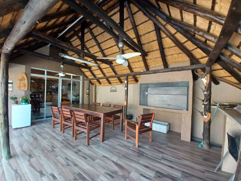 Spacious covered patio featuring a traditional thatched roof, exposed timber beams, built-in braai, and tiled flooring, perfect for outdoor entertaining.