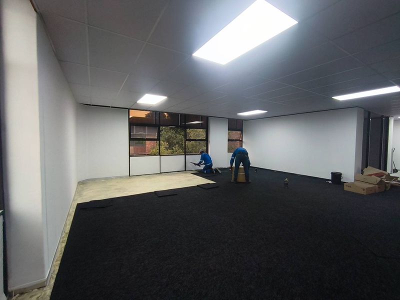 An open-plan office space currently undergoing carpet tile installation, featuring a suspended ceiling with recessed lighting and large windows.