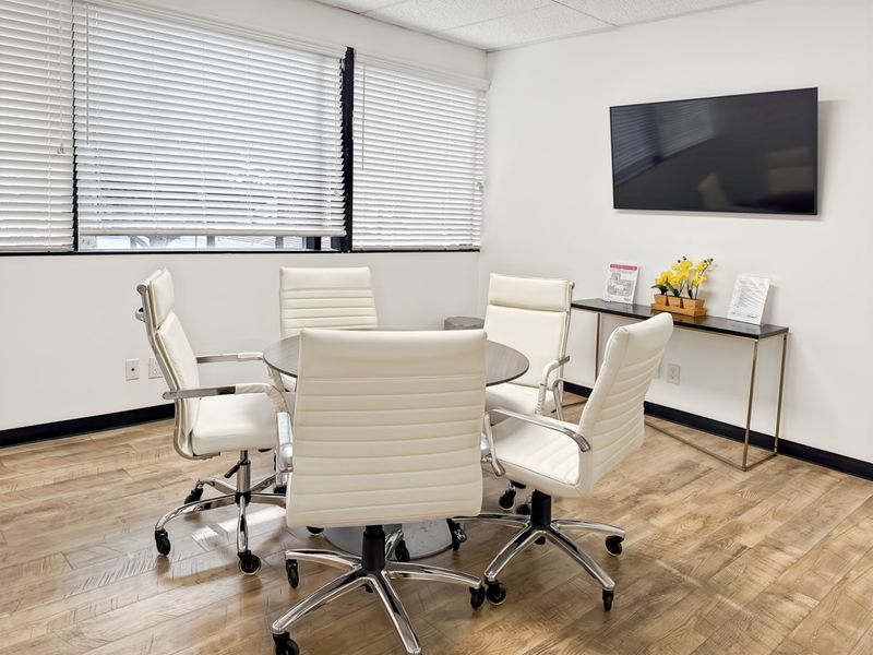 A bright and professional office meeting room featuring wood-look flooring, large windows with venetian blinds, and a wall-mounted display screen.