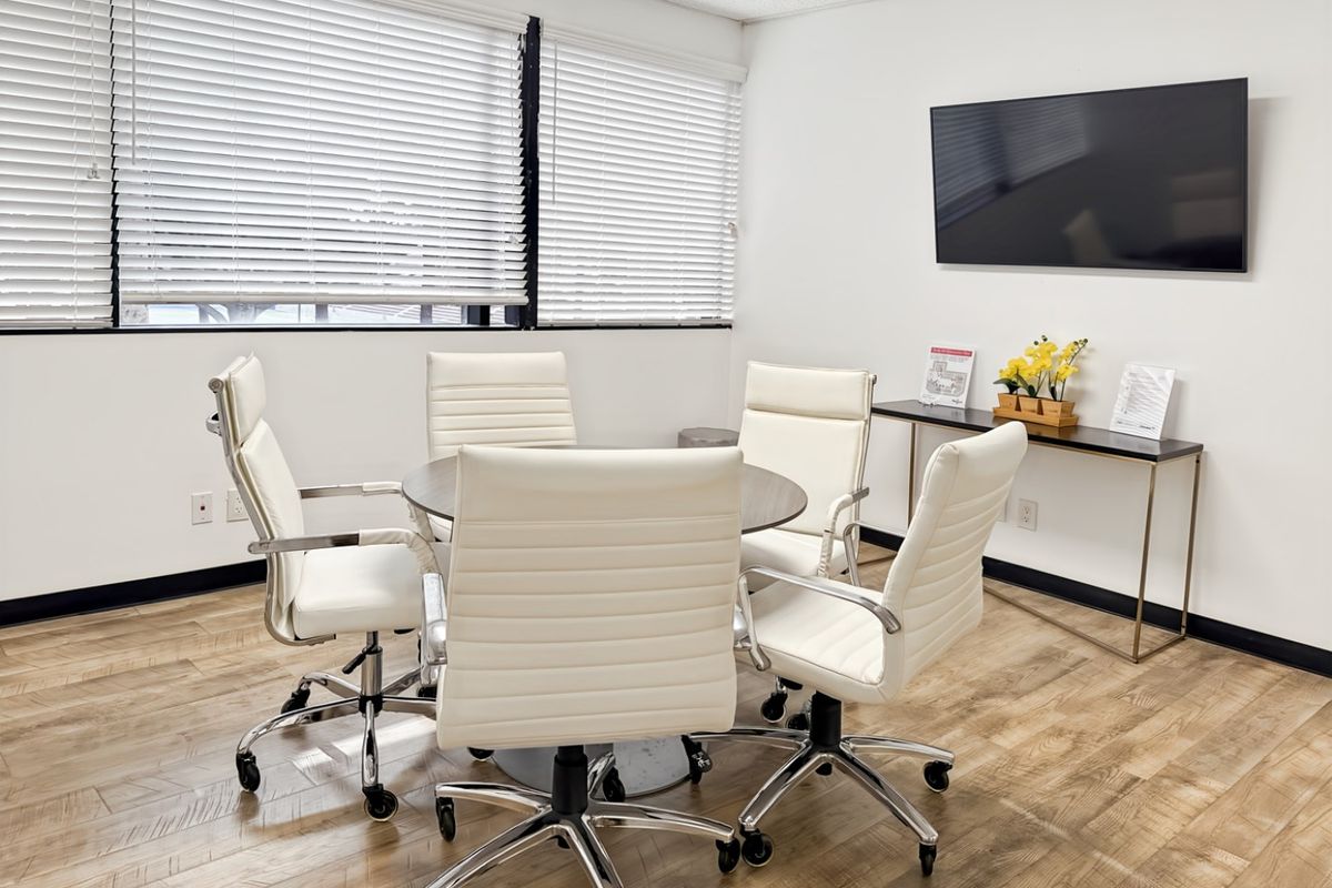 A bright and professional office meeting room featuring wood-look flooring, large windows with venetian blinds, and a wall-mounted display screen.
