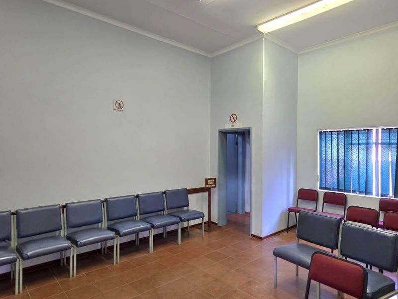 A functional office waiting area featuring tiled flooring, fluorescent lighting, and vertical window blinds, providing a practical space for visitors.