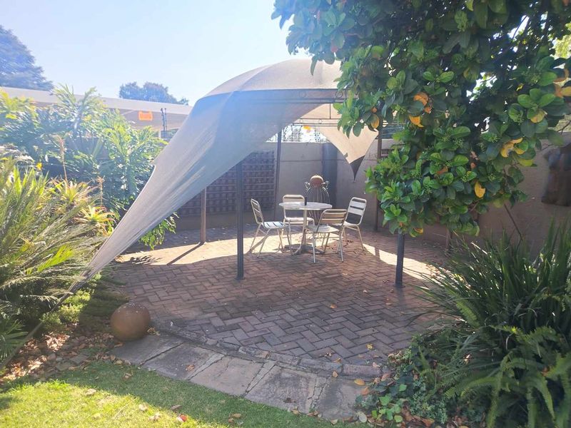 A paved outdoor patio area featuring a shade sail, enclosed by a boundary wall and surrounded by established garden landscaping.