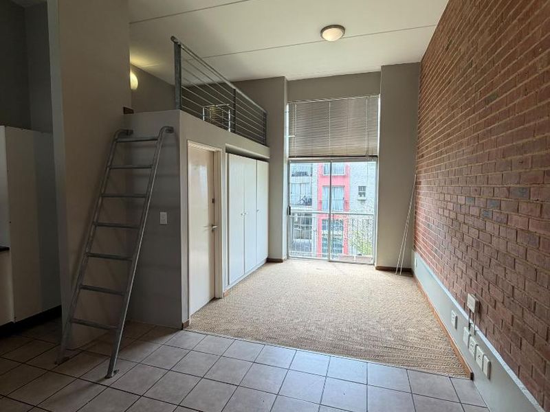 Open-plan apartment living area featuring an exposed brick wall, mezzanine level with ladder access, built-in storage, and a sliding door leading to a balcony.