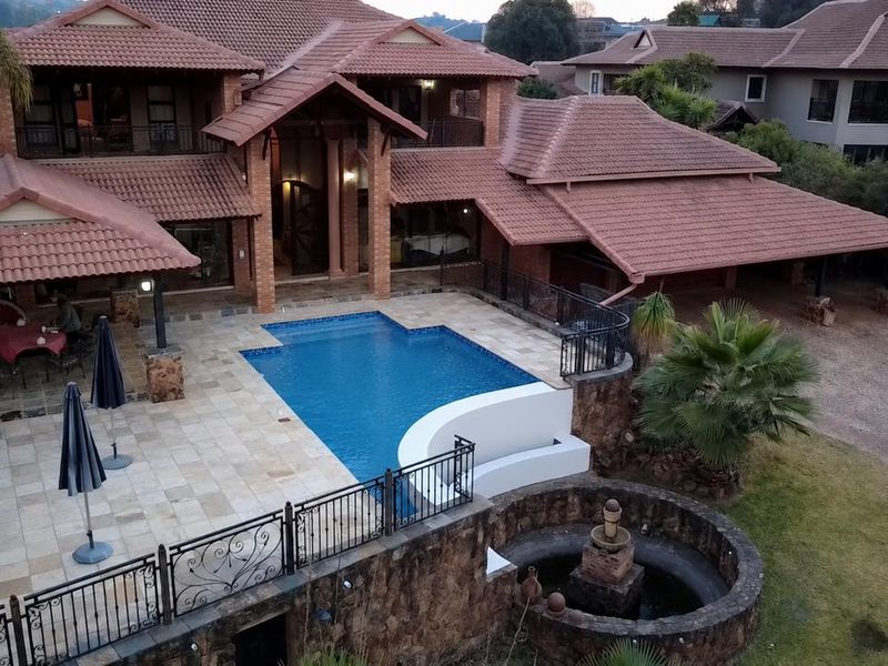 Aerial view of a large residential property featuring a swimming pool, expansive tiled patio, stone water feature, and multi-level brick architecture.
