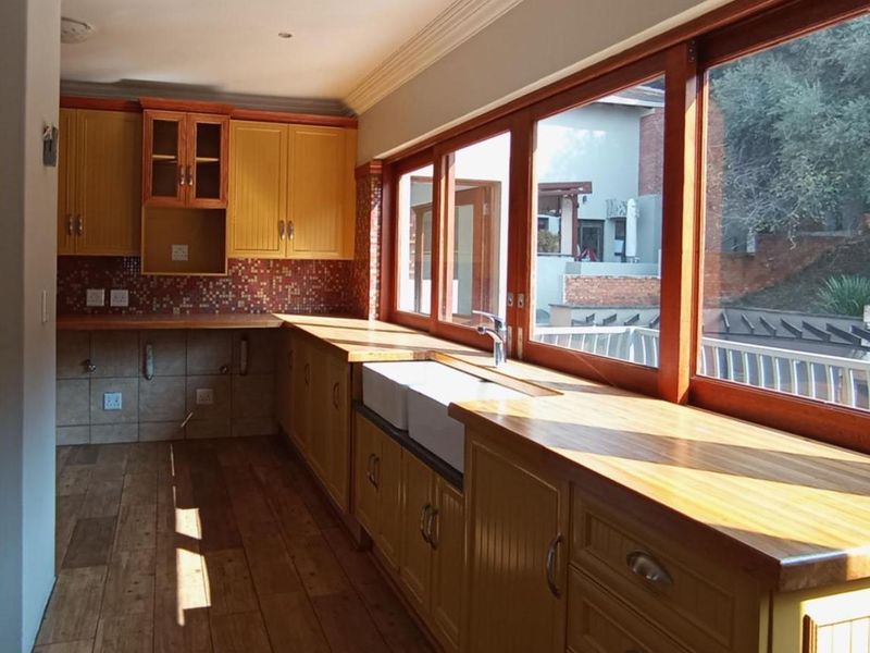 A bright, empty kitchen featuring wooden cabinetry, a classic butler sink, timber-framed windows, and warm wood-look flooring with a mosaic tile backsplash.