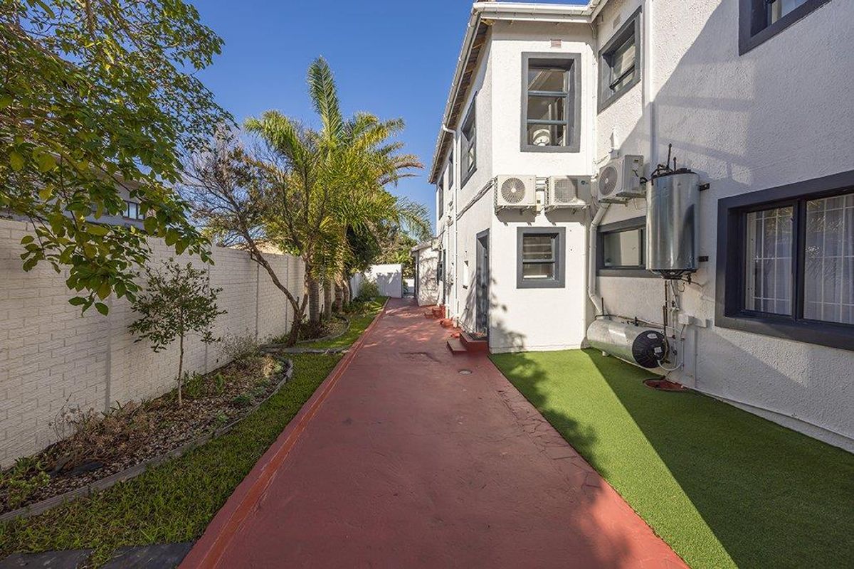 Rear exterior view showing a paved walkway, artificial turf, boundary wall, and external utility installations including air conditioning units and a water heater.