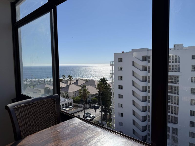 A clear view of the ocean and surrounding neighborhood captured through large, black-framed windows from an elevated apartment position.