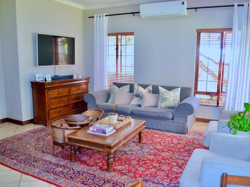 A comfortable living room featuring tiled flooring, a wall-mounted air conditioning unit, and large windows with timber venetian blinds.