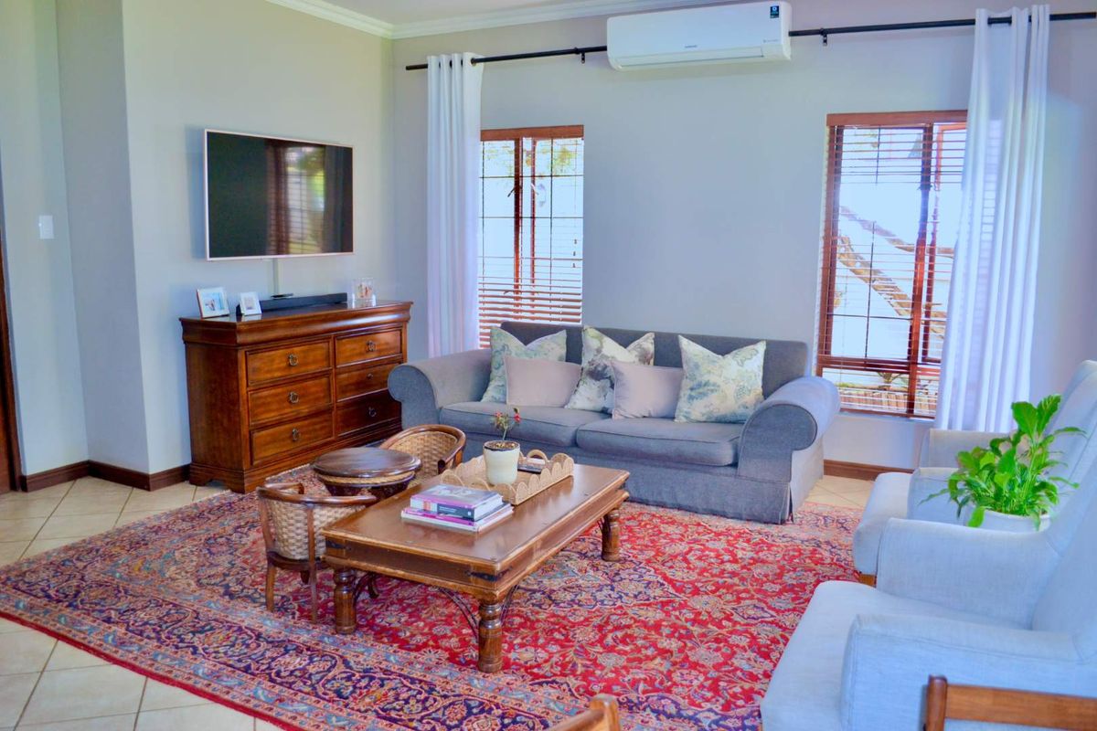 A comfortable living room featuring tiled flooring, a wall-mounted air conditioning unit, and large windows with timber venetian blinds.