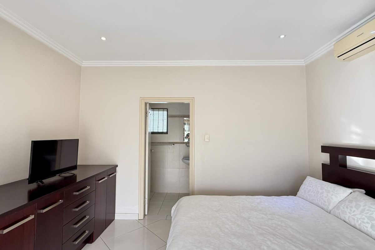 A well-maintained bedroom featuring tiled flooring, crown molding, recessed lighting, and a wall-mounted air conditioning unit, with an adjoining en-suite bathroom.