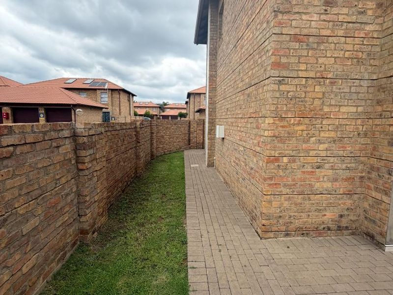 A side exterior view of the property featuring a face brick facade, a paved walkway, and a surrounding boundary wall with a small lawn area.
