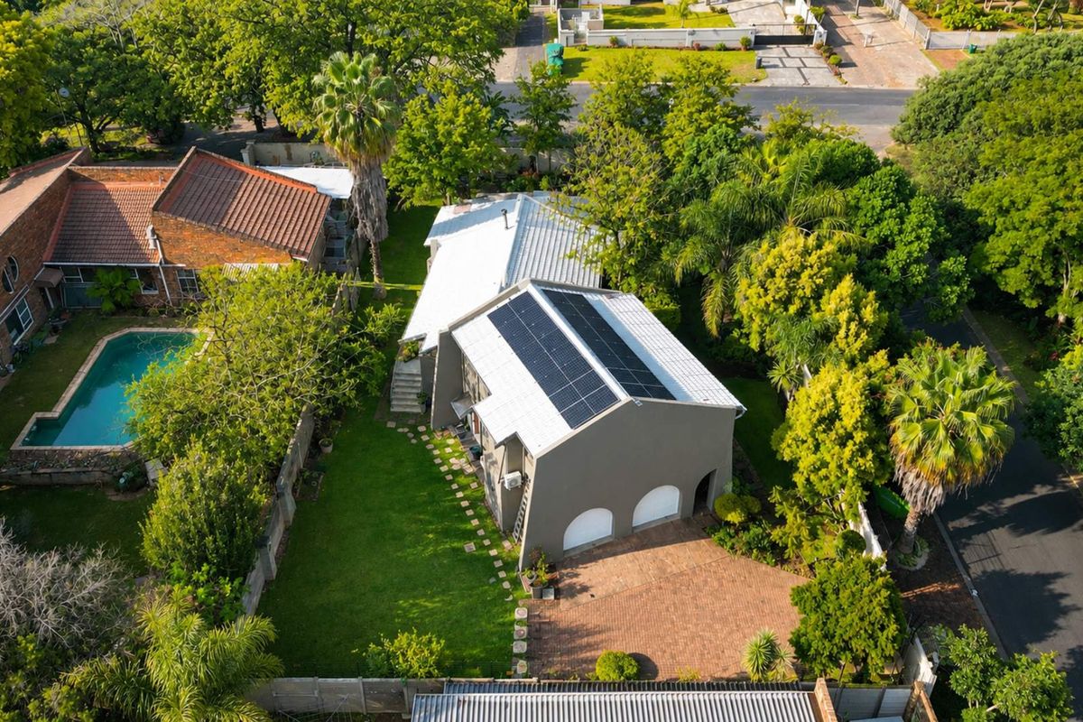 Aerial view of the property featuring a metal roof with solar panels, a paved driveway, double garage, and a well-maintained garden.