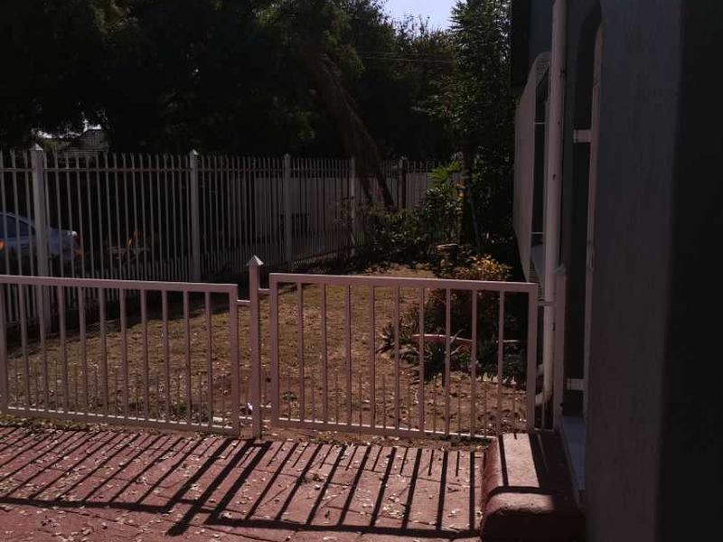 A rear exterior view featuring a paved patio area, metal perimeter fencing, and a small garden space adjacent to the house structure.